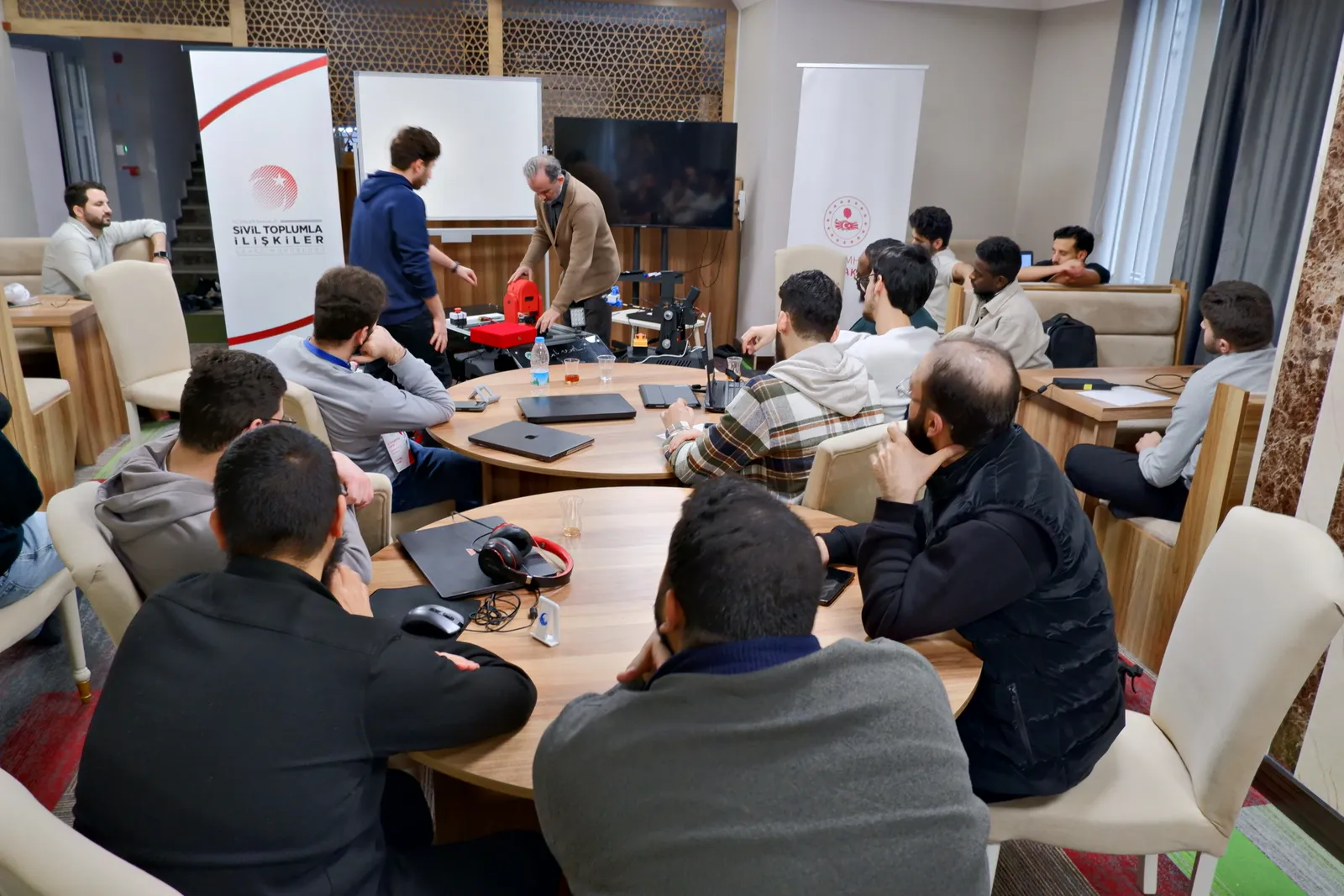 Katılımcılar yuvarlak masalarda, masada robotlar