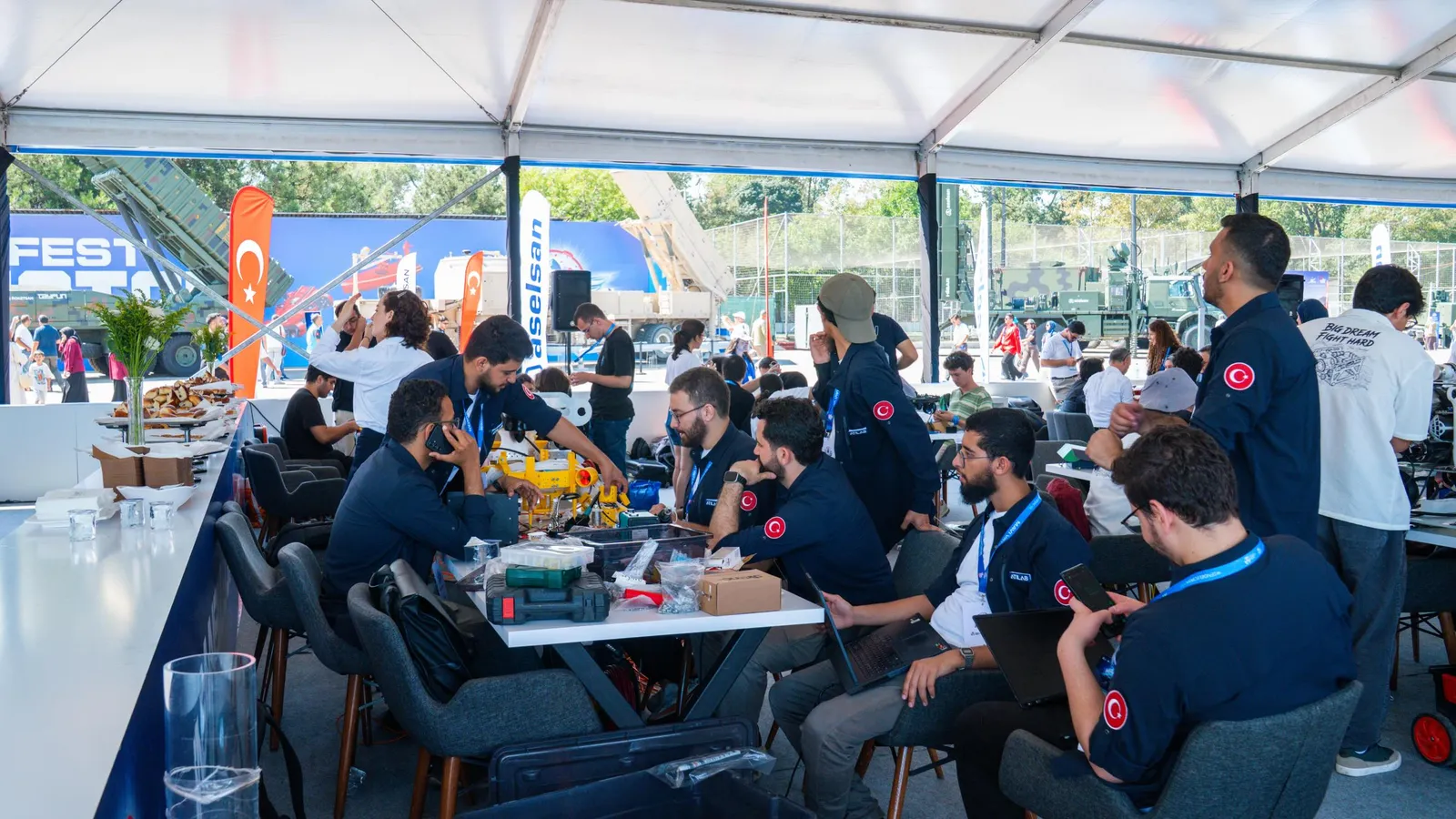 Takım üyeleri çadırda masaların çevresinde projeleri üzerinde çalışıyor
