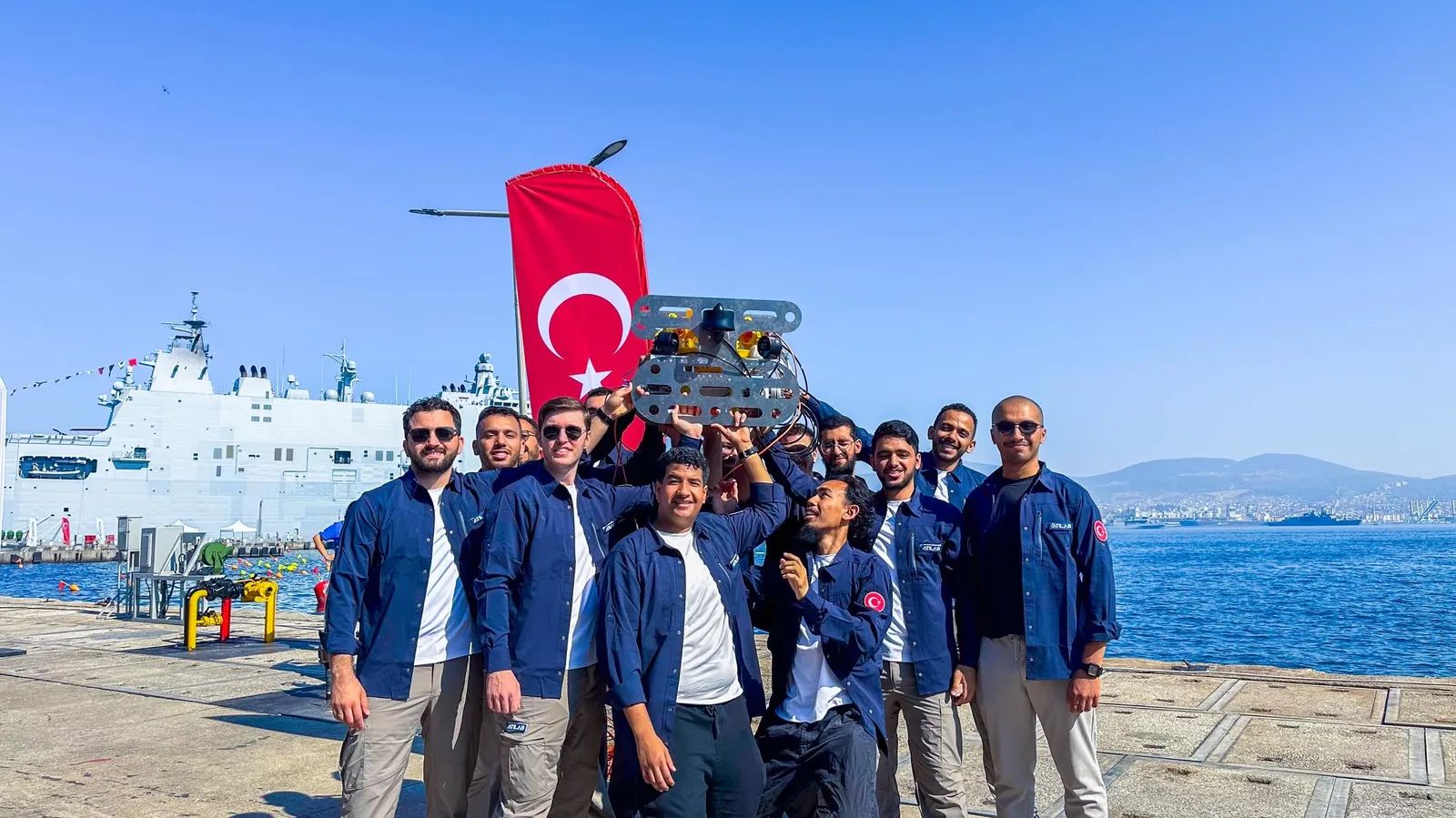 Takım deniz kıyısında su altı aracını havaya kaldırıyor, arkada bir gemi